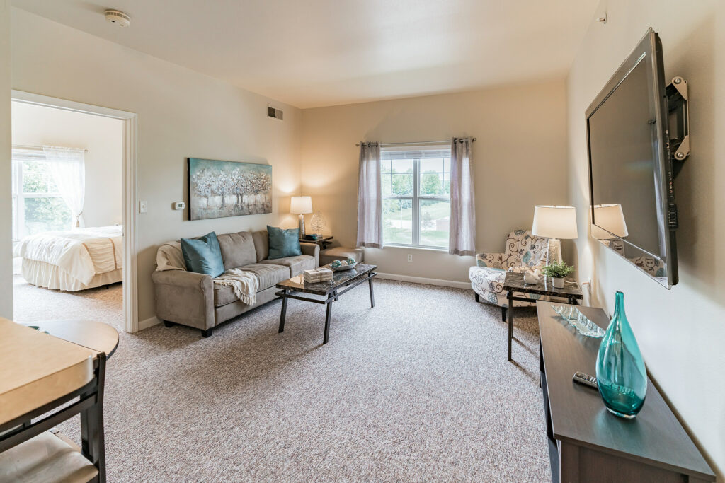 A living room with a gray couch and blue throw pillows on it, a coffee table in front of it, a sofa chair, and a wall-mounted TV above a TV stand at Town Village Sterling Heights, a senior living community near Detroit.