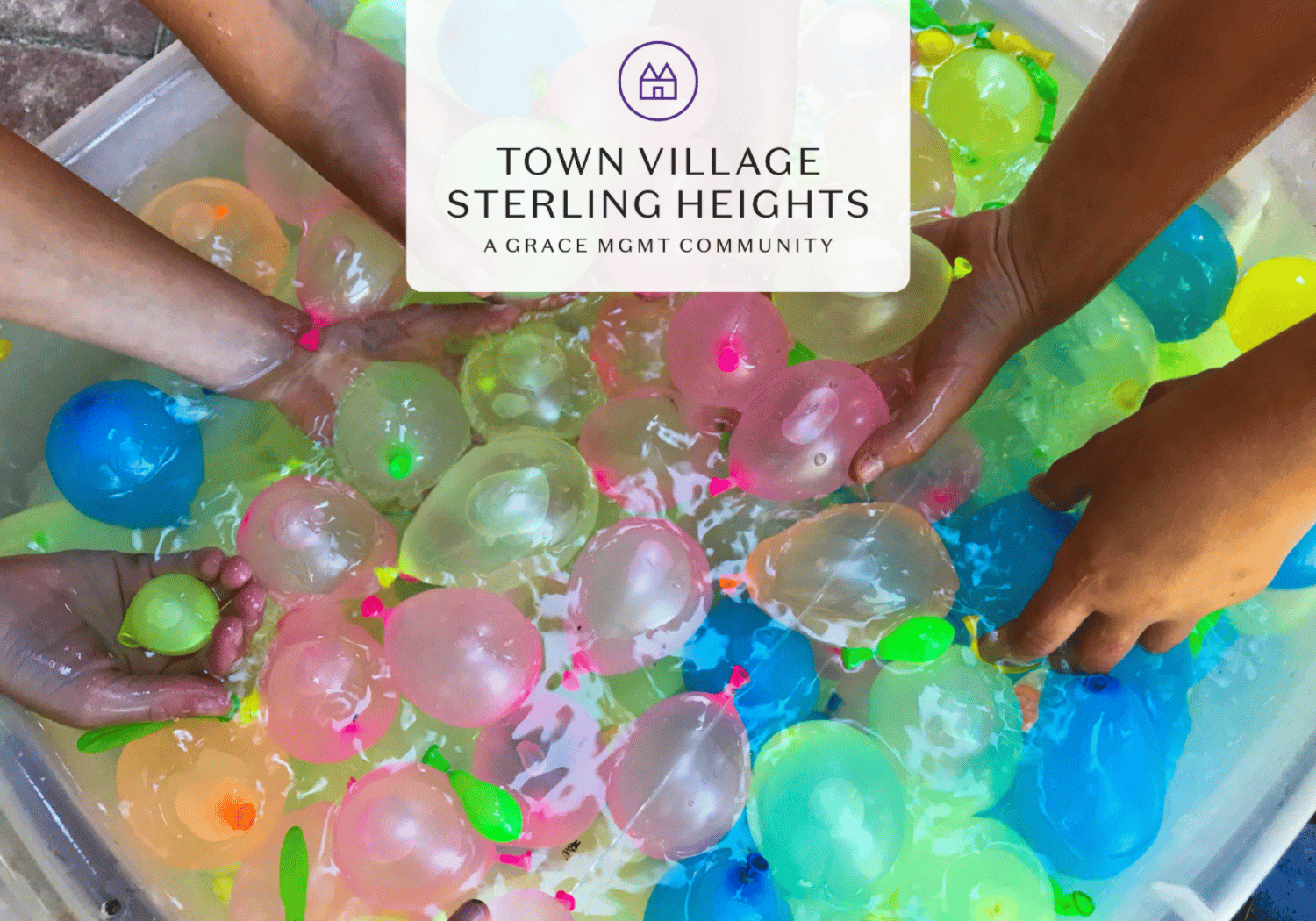 Hands reach into a tub full of colorful water balloons for a community activity, with a white logo banner in the center reading Town Village Sterling Heights.