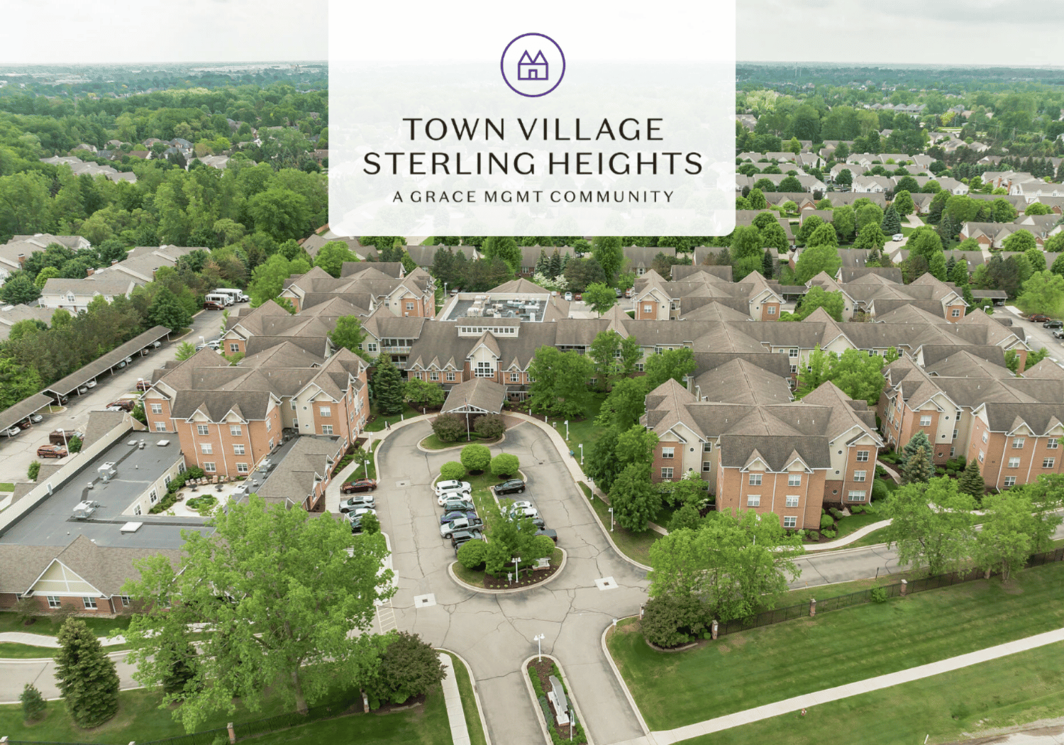 Aerial view of a suburban townhouse community with a central roundabout and parking lots, overlaid by a translucent banner that reads 'Town Village Sterling Heights' and 'A Grace Mgmt Community'.
